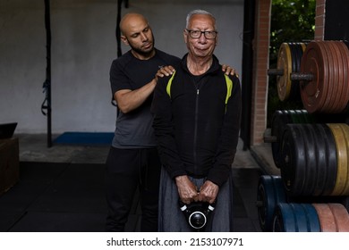 Latin American Senior Male With His Personal Trainer Do Rehabilitation And Therapy Exercises Holding A Kettlebell With Both Hands In The Gym. Healthy Lifestyle Concept.