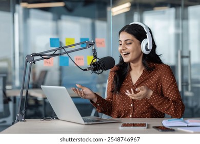 Latin American woman businesswoman recording podcast with microphone and laptop, wearing headphones. She's in a modern workspace, expressing enthusiasm. Smartphone visible on desk. - Powered by Shutterstock