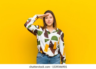 Latin American Woman Greeting The Camera With A Military Salute In An Act Of Honor And Patriotism, Showing Respect Isolated Against Yellow Wall