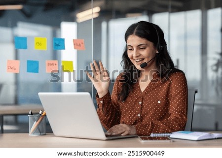 Latin American woman with headset phone using laptop for video call, female online customer support worker waving hand greeting customer, online meeting with colleagues remotely.