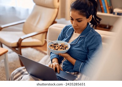 Latin American woman surfing the net on laptop while eating breakfast cereal in the living room.  - Powered by Shutterstock