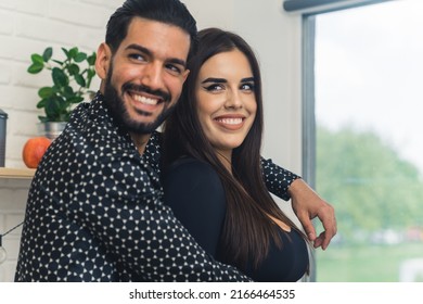 Latin Bearded Dark-haired Man In His 30s Hugging A Pretty Brunette Millenial Woman, Looking Away, Standing In A Modern Kitchen. High Quality Photo