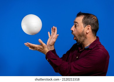 Latin Bearded Man Dressed In A Purple Shirt With A Cork Ball Isolated On Blue Studio Background, He Is Throwing The Ball Imitating Son Goku From Dragon Ball One Of His Youth Heroes.