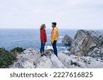 latin boy and caucasian girl in red jacket and yellow jeans standing in profile facing each other looking at each other and smiling on the big rocks, cabo de penas, spain