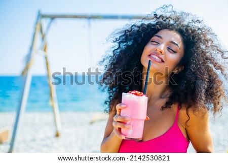 latin brazilian woman with curly long hairstyle and pink bikini drinking strawberry smoothie in beach on the swing