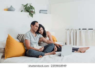 Latin couple watching a movie on laptop or computer lying on the bed at home in Mexico Latin America, hispanic people eating popcorn on bedroom - Powered by Shutterstock