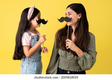 Latin excited mother and little girl laughing while playing with fake mustaches in front of a bright studio background