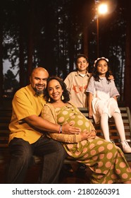 Latin Family With Pregnant Mom, Dad, Son And Daughter Sitting On Park Bench In Pine Trees At Night Looking At The Camera