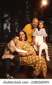 Latin Family With Pregnant Mom, Dad, Son And Daughter Sitting On Park Bench In Pine Trees At Night Looking At The Camera