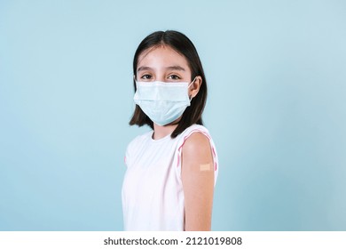 Latin Girl Child Wearing Face Mask After Getting A Vaccine Protection The Coronavirus And Showing Arm With Bandage After Receiving Vaccination On Blue Background