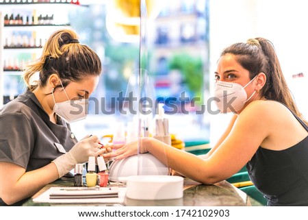 Latin girl with face mask painting colored nails to the brunette client. Reopening after the corod-19 pandemic. Manicure and Pedicure Salon. Coronavirus