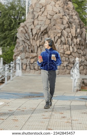 Latin girl with headphones and a disposable cup of coffee walking through a public park.