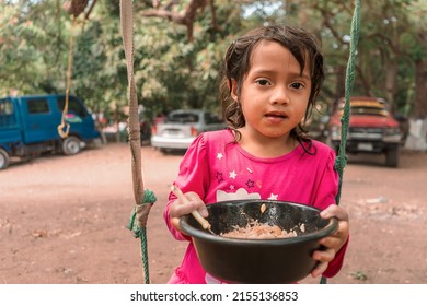 Latin Girl Sitting On A Swing Eating From A Plastic Bowl. Concept Of Childhood, Nutrition And Food In Rural Areas Of Nicaragua Latin America