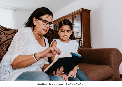 Latin Grandmother With Granddaughter Sitting On Couch At Home And Reading Book In Mexico City