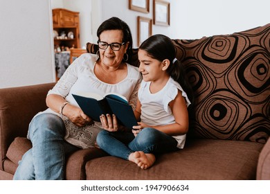 Latin Grandmother With Granddaughter Sitting On Couch At Home And Reading Book In Mexico City