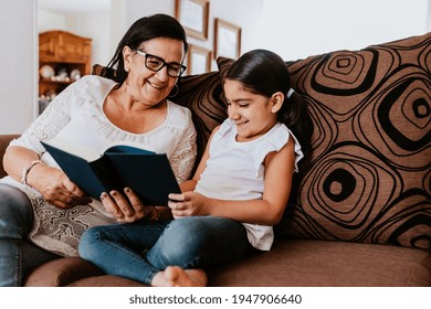 Latin Grandmother With Granddaughter Sitting On Couch At Home And Reading Book In Mexico City