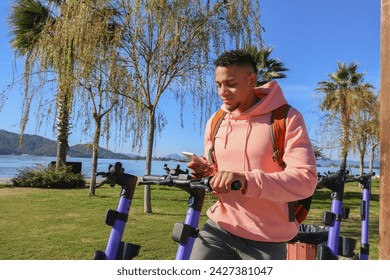 Latin guy in a pink hoodie scanning a QR code to rent an e-scooter in the park. Mixed race young man about to unlock and ride an electronic scooter. Close up, copy space, background. - Powered by Shutterstock