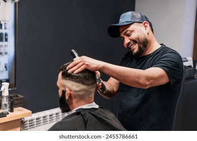 A Latin hairdresser cuts the hair of a man with a beard. The barber is wearing a baseball cap and is smiling and concentrating on his task. - Powered by Shutterstock