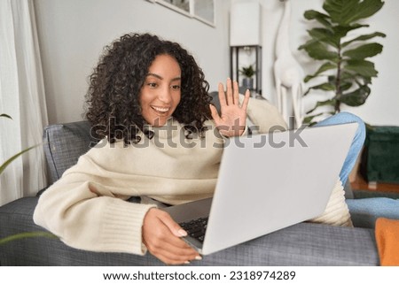 Latin happy smiling curly young woman sitting on couch using laptop at home waving hand, having video call communicating in online dating videocall chat, looking at computer relaxing at home.