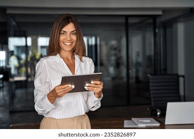 Latin Hispanic 40s years mature business woman looking at camera. smiling European businesswoman CEO holding digital tablet using fintech tab online application standing in modern office, copy space - Shutterstock ID 2352826897