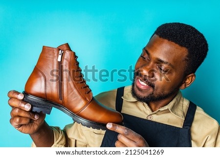 latin hispanic man in black apron showing brown leather shoes in blue studio background