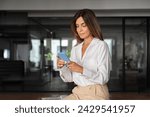 Latin Hispanic mature adult professional business woman working on mobile cellphone European businesswoman CEO holding digital smartphone using fintech tab application standing in office, copy space