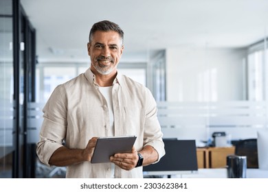 Latin Hispanic mature adult professional business man looking at camera and smiling. Indian senior businessman CEO holding digital tablet using fintech tab application standing inside company office. - Powered by Shutterstock