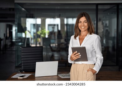 Latin Hispanic mature adult professional business woman looking at camera and smiling. European businesswoman CEO holding digital tablet using fintech tab application standing at workplace in office. - Shutterstock ID 2343004285