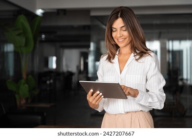 Latin Hispanic mature adult professional business woman looking at online trade app. European businesswoman CEO holding digital tablet using fintech tab application standing at workplace in office.