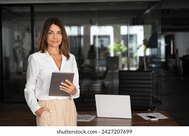 Latin Hispanic mature adult professional business woman looking at camera and smiling. European businesswoman CEO holding digital tablet using fintech tab application standing at workplace in office.