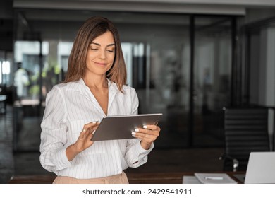 Latin Hispanic mature adult professional business woman looking at online trade app. European businesswoman CEO holding digital tablet using fintech tab application standing at workplace in office.