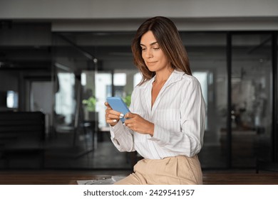 Latin Hispanic mature adult professional business woman working on mobile cellphone European businesswoman CEO holding digital smartphone using fintech tab application standing in office, copy space