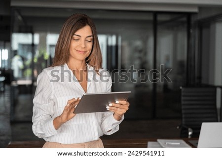 Latin Hispanic mature adult professional business woman looking at online trade app. European businesswoman CEO holding digital tablet using fintech tab application standing at workplace in office.