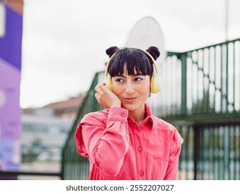 Latin hispanic woman whit  yellow headphones look away in the city while enjoy music whit buns hairstyle. Wear pink jacket. Technology lifestyle. Ecuador beauty - Powered by Shutterstock