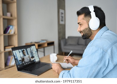 Latin Indian Adult Student Wearing Headset Having Virtual Meeting Online Call Training Educational Webinar Chatting With Teacher At Home Office Writing Notes. Video E Learning Conference Call On Pc.