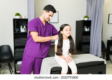 Latin Male Physiotherapist Giving A Shoulder Massage To A Little Girl Patient. Cheerful Elementary Kid Relaxing 