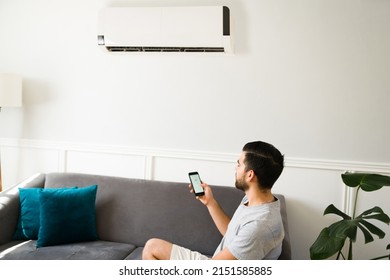 Latin Man Checking The Weather App On His Smartphone During The Summer While Using The Air Conditioner