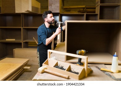 Latin Man In His 30s Doing Woodworking In A Big Woodshop. Male Carpenter Hammering A Screw In A Piece Of Furniture