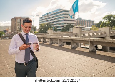 Latin man with a tie walking through the financial center of Guatemala with his cell phone and a coffee cup in his hands. Buildings and flag of Guatemala in the background.