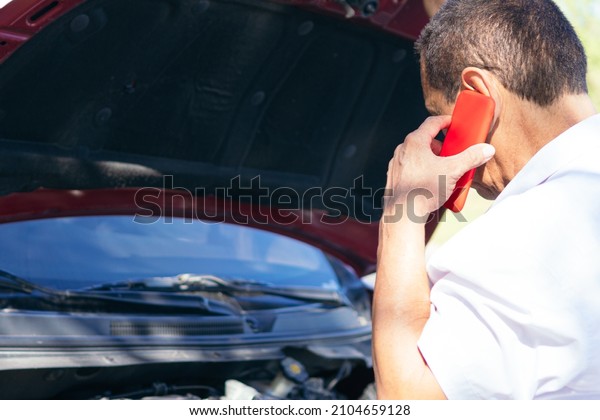Latin Man trying to call\
service, standing in front of the open hood of a broken down car\
outside highway