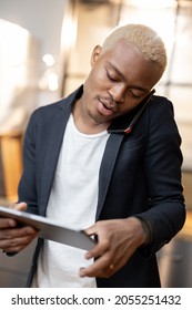 Latin Man Using Digital Tablet While Talk On Smartphone. Concept Of Modern Domestic Lifestyle. Young Focused Guy Work Remotely From Home. Interior Of Kitchen In Modern Apartment