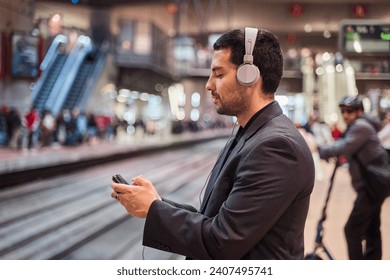 latin man waiting for the train while using his smart phone with headphones on playing games or watching a video