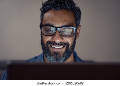 Latin man working on computer at night in dark office. Portrait of handsome hispanic businessman wearing glasses working with laptop. Closeup face of indian man using digital tablet in the evening.