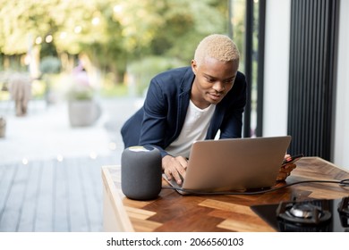 Latin Man Works On A Laptop Computer At Thhe Kitchen Table With Smart Speaker. Controlling Smart Home Devices Remotely With A Voice Command. Concept Of Smart Home. Idea Of Audio Technology