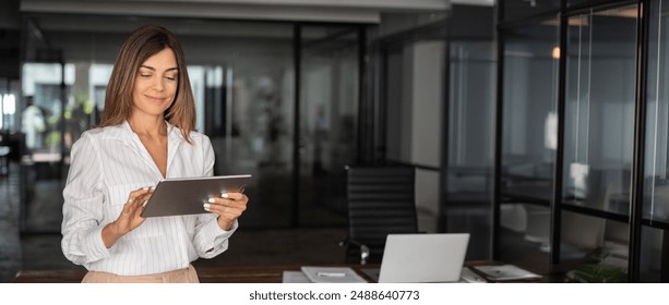 Latin mature adult professional business woman looking at online trade app. European businesswoman CEO holding digital tablet using tab application standing at workplace in office. Banner, copy space - Powered by Shutterstock