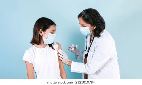 Latin Medical Woman Doctor Or Nurse Giving Coronavirus Vaccine Shot To Hispanic Child Girl Over Blue Studio Background