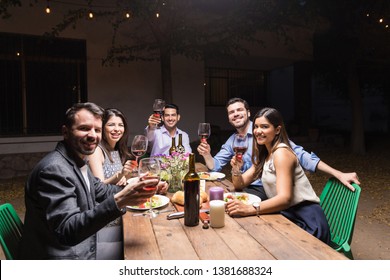 Latin Men And Women Raising Red Wine Glasses During Dinner Party In Yard