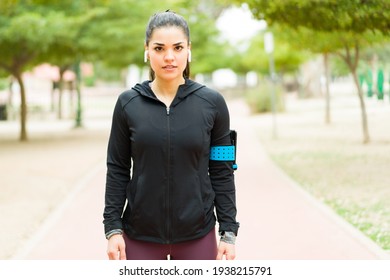 Latin Pretty Woman In Activewear Making Eye Contact With A Serious Expression. Fit Hispanic Woman Ready To Start Running Outdoors With Headphones And A Smartphone In Her Arm 