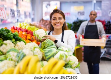 Latin saleswoman in black apron and her assistant with box of vegetables in background