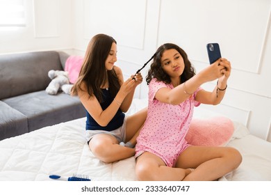 Latin teen girl taking a selfie with her best friend while doing braids in their hair during their fun sleepover in the bedroom - Powered by Shutterstock